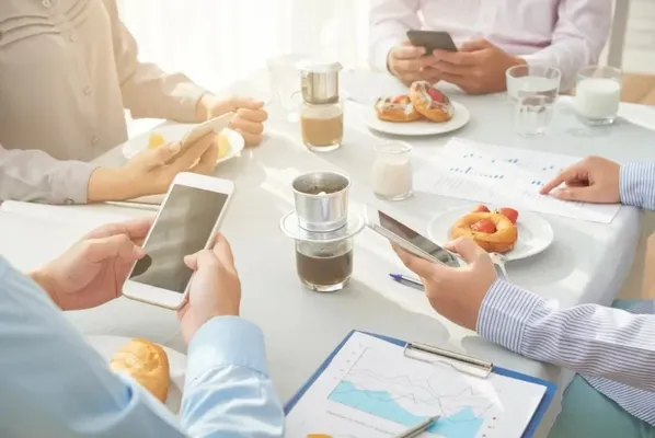 Hay 4 trabajadores en una mesa, están con sus teléfonos celulares en las manos, toman un coffee break ejecutivo, enfrente hay platos con pan de dulce y sus respectivas tazas de café, en la mesa se ve que hay reportes del trabajo y tiene un mantel blanco,hay vasos con leche en la esquina de donde esta cada persona