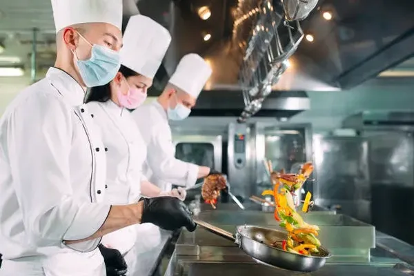 Varios chefs que están frente a las parrillas haciendo comida para el servicio de catering, a la izquierda hay un hombre, enmedio una mujer y en la derecha hay otro hombre, los tres estan usando un gorro, camisas blancas y guantes negros, los tres usan cubrebocas, los hombres traen cubrebocas azules y la mujer un cubrebocas rosa, el hombre de la izquierda esta volteando unas verduras en el sartén