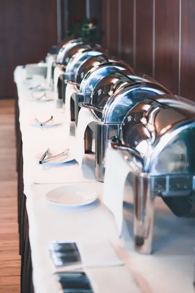 Mesa con un mantel blanco, que tiene varias cacerolas con comida, porque se va a llevar a cabo un buffet para eventos, las charolas tienen unos manteles blancos que sobresalen, hay platos enfrente y cubiertos, atrás hay una pared de madera.