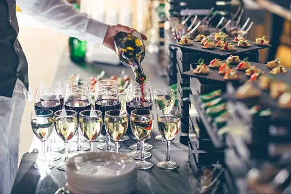 Hay varias copas de vino que están siendo llenadas para el servicio de catering, las copas tienen con vino blanco y vino tinto, la persona encargada esta sirviendo el vino tinto, atrás hay varios platos de comida con canapes, los platos son de color café, también hay platos blancos para poner la comida