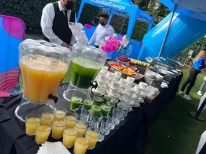 Servicio de catering, donde hay muchos vasos de agua, una jarra dispensadora con agua de color amarilla, hay vasos que ya están llenos y al lado hay una jarra con agua de color verde, los vasos apenas se estan llenando, al lado de las jarras hay muchas tazas sobre platos, las tazas y platos son blancos, al lado hay una cacerola con comida, hay platos apilados, la mesa tiene un mantel negro, atrás hay un inflable azúl y hay dos meseros con camisa blanca