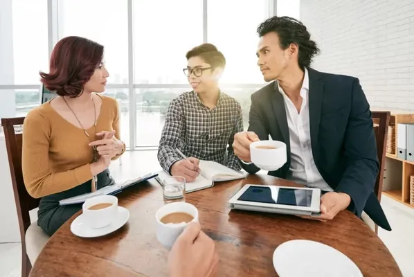 Hay 4 personas que están tomando un coffee break ejecutivo, están alrededor de una mesa de madera, se visualizan dos hombres, uno de ellos tiene un saco negro con un ¡Pad en la mano y una taza de café en la otra, al lado hay un hombre con camisa y lentes que escribe en un cuaderno, ambos están viendo a una mujer que tiene una libreta en las piernas y una taza de café enfrente, enfrente de ellos se visualiza una mano extendida sosteniendo un café