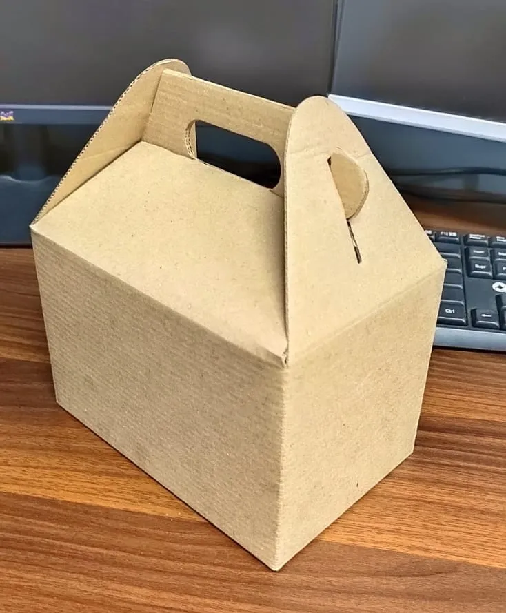 Catering box lunch, es una caja con agarraderas, es de cartón, cerrada, puesta sobre una mesa de madera