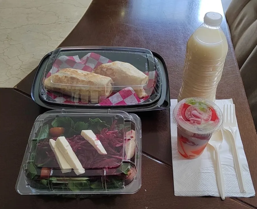 Dos charolas de comida individuales, hay una botella de agua, esta puesta sobre una mesa de madera, está lista para box lunch a domicilio