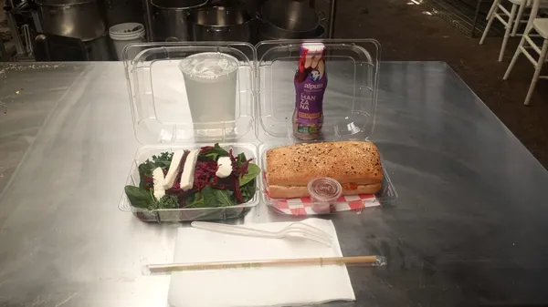 Sobre una mesa hay un box lunch, hay una charola con comida y dos bebidas