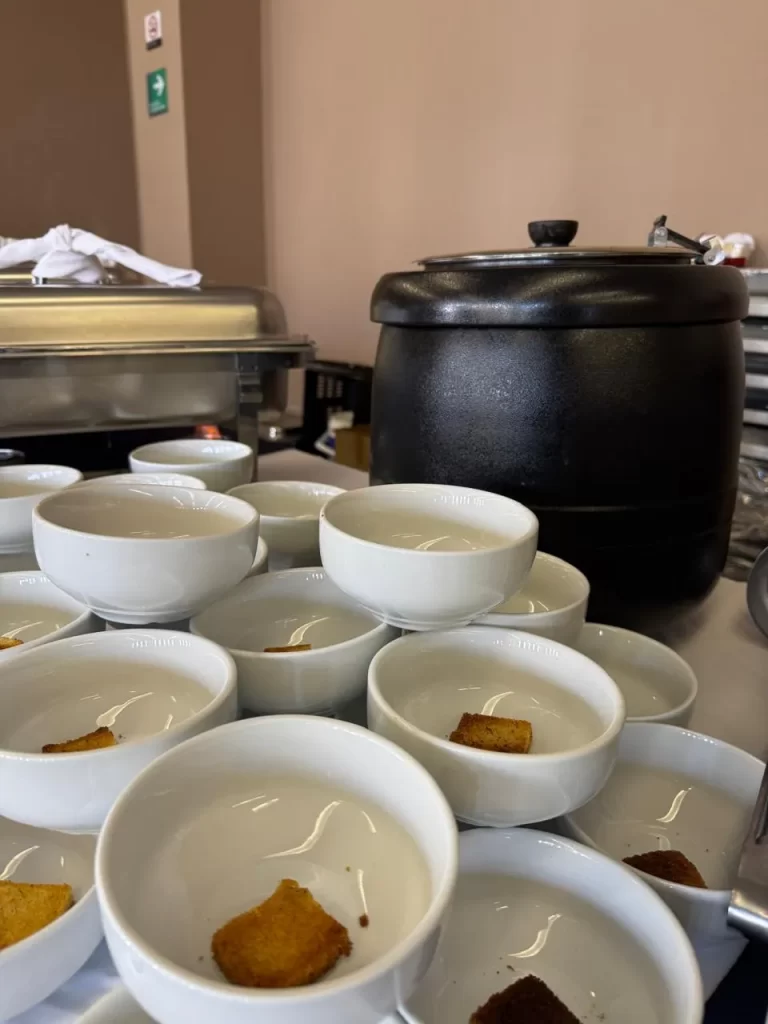 Esta imagen captura una estación de buffet preparada para servir sopa o crema. Al fondo, se vislumbra un calentador de alimentos lo que sugiere un buffet bien organizado y listo