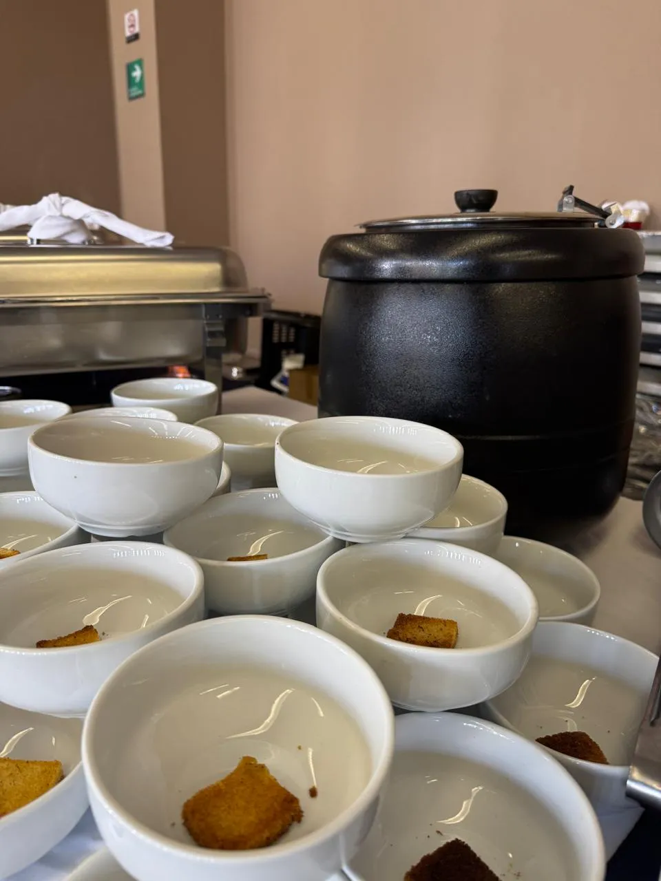 Esta imagen captura una estación de buffet preparada para servir sopa o crema. Al fondo, se vislumbra un calentador de alimentos lo que sugiere un buffet bien organizado y listo