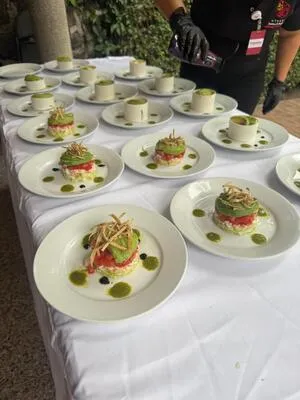 Fila de platos individuales listos para los comensales, con una torre de aguacate y postres en el fondo, preparada por el personal de servicio de comida para eventos.