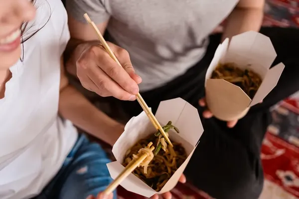 Dos personas comparten comida de sus respectivos box lunch cdmx con palillos, sentadas sobre una alfombra.