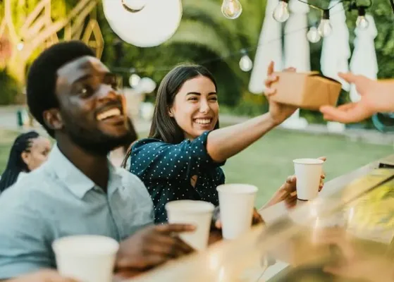 Dos personas sonrientes reciben sus box lunch cdmx en un evento al aire libre, con vasos en la mano.