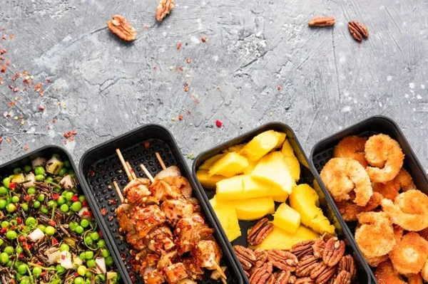 Una vista aérea de cuatro box lunch cdmx abiertos que contienen camarones, pollo, ensalada de arroz y mango con nueces.